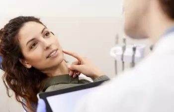 A dentist and a concerned female patient sitting in a dental chair and pointing at her lower jaw.