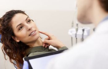 A dentist and a concerned female patient sitting in a dental chair and pointing at her lower jaw.